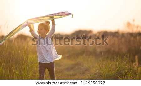 A girl runs into a field with a kite, learns to launch it. Outdoor entertainment in summer, nature and fresh air. Childhood, freedom and carelessness. A child with wings is a dream and a hope.
