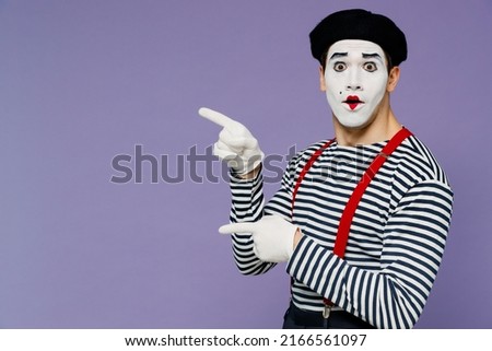 Surprised ecstatic young mime man with white face mask wears striped shirt beret pointing aside on workspace area copy space mock up isolated on plain pastel light violet background studio portrait