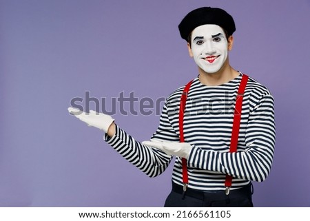 Happy smiling young mime man with white face mask wears striped shirt beret pointing aside on empty palm with copy space place mock up isolated on plain pastel light violet background studio portrait Royalty-Free Stock Photo #2166561105