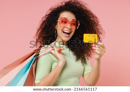 Side view vivid young curly latin woman 20s wears mint t-shirt sunglasses hold package bags with purchases after shopping credit bank card isolated plain pastel light pink background studio portrait