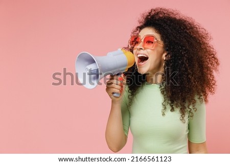 Exultant happy vivid young curly latin woman 20s wears mint t-shirt sunglasses hold scream in megaphone announces discounts sale Hurry up isolated on plain pastel light pink background studio portrait Royalty-Free Stock Photo #2166561123