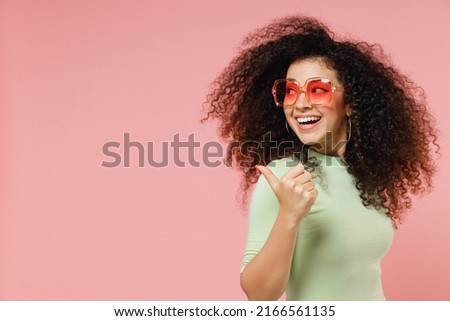 Fun young curly latin woman 20s wears mint t-shirt sunglasses pointing pointing thumb finger behind on workspace area copy space mock up isolated on plain pastel light pink background studio portrait