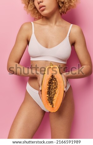 Cropped shot of unknown woman dressed in underwear holds half of papaya fruit uses tropical fruits for making natural cosmetic products isolated over pink background shows beauty of her body