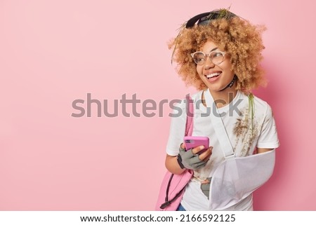 Happy active woman cyclist wears sling on arm looks cheerful away wears dirty t shirt and helmet uses smartphone concentrated away isolated over pink background empty space for your advertisement