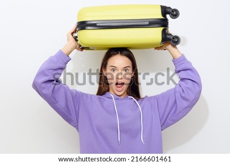 a surprised woman standing on a white background in a purple tracksuit and a yellow cap lifted a suitcase over her head with her mouth and eyes wide open