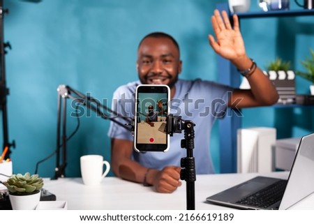 Closeup of live vlog setup with smarthone on stand filming content creator waving hand to followers in online morning show. Selective focus on vlogger saying hello at audience sitting at desk.