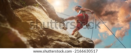 Muscular climber man in protective helmet abseiling from cliff rock wall using rope Belay device and climbing harness on evening sunset sky background. Active extreme sports time spending concept. Royalty-Free Stock Photo #2166630517