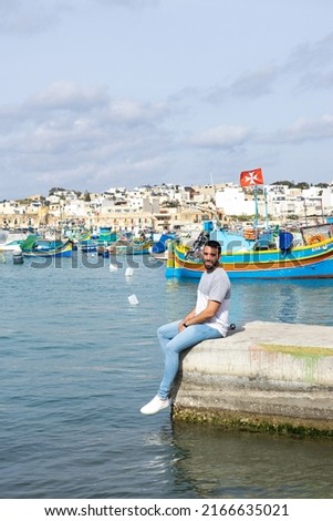 Young man seated and in the background the sea full of colorful boats in Marsaxlokk