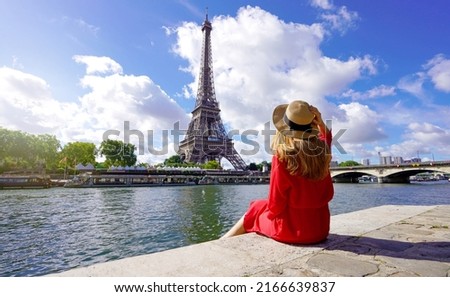 Young traveler woman in red dress and hat sitting on the quay of Seine River looking at Eiffel Tower, famous landmark and travel destination in Paris. Royalty-Free Stock Photo #2166639837