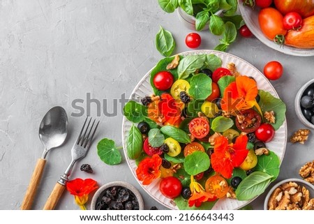 Nasturtium salad with vegetables, olives, nuts and raisins on gray concrete background with cutlery. Top view. Healthy diet food. Flat lay