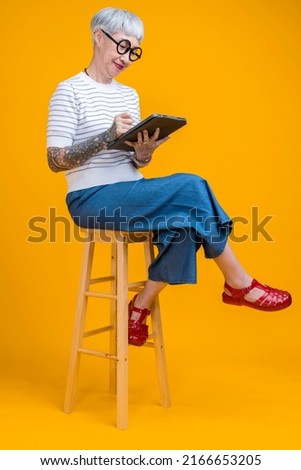 smiling retired old senior asian female with tattoo relax on comfy stool use modern digital touchpad tablet device read ebook from screen scroll social media web pages isolate yellow background Royalty-Free Stock Photo #2166653205