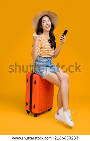 ready to travel ,asian smiling cheerful female woman hand using smartphone check flight schedule pull luggage bag prepare to new abroad journey travel studio shot on yellow background 