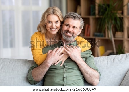 Happy Middle Aged Couple. Romantic Man And Woman Hugging At Home, Portrait Of Loving Mature Spouses Embracing And Smiling At Camera While Relaxing Together On Couch In Living Room, Closeup