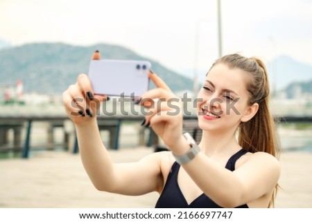 Cute caucasian woman wearing sportive clothes on city park, outdoors taking horizontal photo with her smart phone. Selective focus on her face.