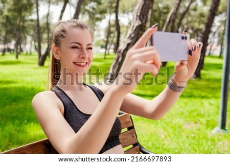 Young brunette girl smiling happy wearing sportive clothes on city park, outdoors taking horizontal photo with her smart phone.