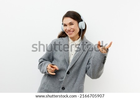 funny funny woman in stylish coat and headphones enjoying music gesturing signs of approval while standing on a light background with empty space