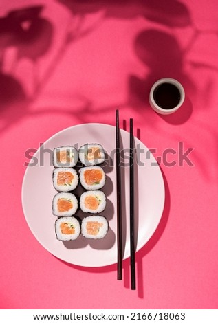 Japanese Sushi roll with salmon, cucumber and soy sauce with chopsticks in the plate on pink background with hard shadow flower leaf. Concept food photography. Top view and copy space.