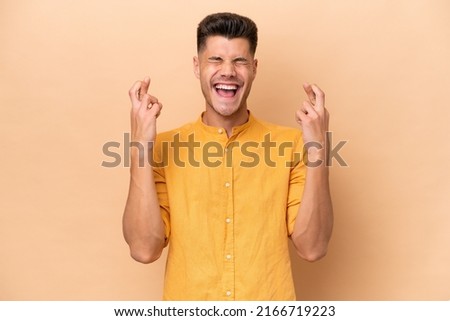 Young caucasian man isolated on beige background with fingers crossing