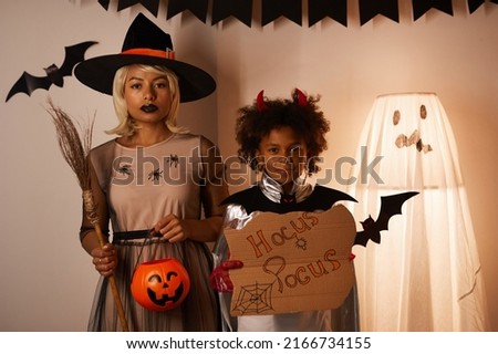 Portrait of serious young black mother in witchs dress and son in devil costume holding cartoon board with hocus pocus inscription, they welcoming to Halloween party