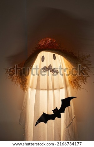 Illuminated lamp covered with sheet placed in dark room, white sheet with drawn ugly face and straw hat