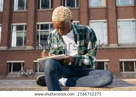 Portrait of pensive man artist wearing casual clothing drawing  something sitting alone on the street. Young stylish writer taking notes holding notepad, listening music, relaxing outdoors