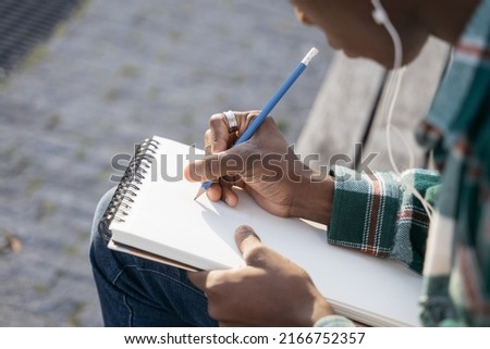Closeup of artist hand drawing, taking notes into paper notepad sitting outdoors, selective focus. Student studying, education concept 
