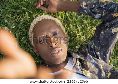 Portrait of young African American guy with stylish hair taking selfie  looking at camera laying on grass. Fashion model posing for pictures outdoors. Beauty concept 