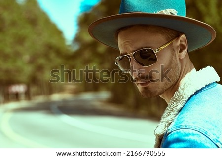 Portrait of a handsome bearded man in denim jacket, a hat and black sunglasses standing by the highway. Denim fashion. Road adventures, hitchhiking, autotrip.  Royalty-Free Stock Photo #2166790555