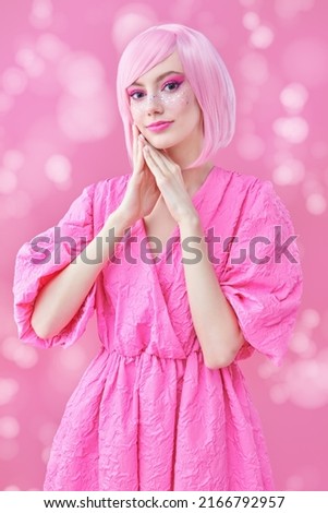 Beauty, fashion. Portrait of a cute teen girl with bright pink makeup and pink hair smiling dreamily and posing in fashionable pink dress. Pink background with lights. 