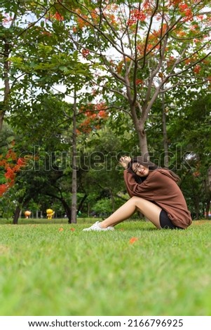Young asian female lady model in her exercise sweater jacket sitting chillingly and calmly on the green grass while enjoying the calming breeze air and sunbath in an outdoor park