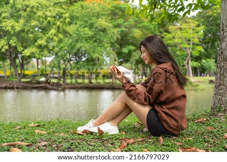 Young asian female lady model in her exercise sweater jacket sitting on green grass reading a generic book while enjoying the calming breeze air and sunbath beside a calm lake in an outdoor park Royalty-Free Stock Photo #2166797029
