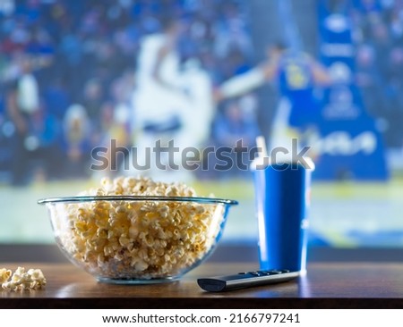 Popcorn in a glass bowl, a drink in a blue plastic glass with a straw and a TV remote control on the table against the background of a large screen of a working TV. Recreation, relaxation, hobby.