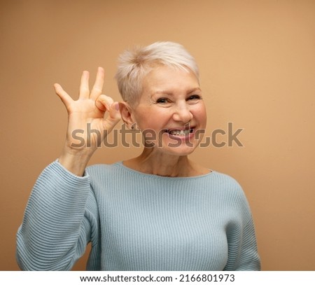 Happy smiling old lady with short hair showing ok sign headshot at studio. Elderly woman giving good advice, offer, help, portrait isolated on beige background.