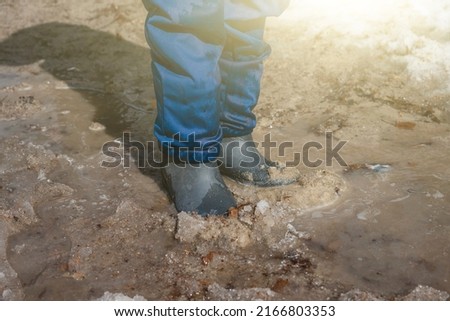 Boy's feet in rubber boots in muddy puddle with sand and snow. Child boy of 6-7 year old walking and playingin wellies in melted earth. Spring came