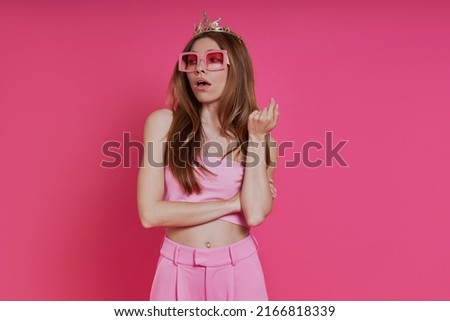 Bossy young woman in funky crown looking displeased while standing against pink background Royalty-Free Stock Photo #2166818339