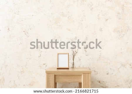 Blank frame and vase with flowers on table near light wall
