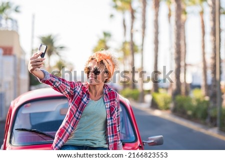 Hippy lifestyle female take selfie picture against her red classic car smiling. Road in background. Concept of people and vehicle transportation. Happy female driver use phone and enjoy trip day
