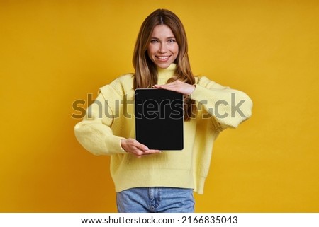 Cheerful young woman showing her digital tablet while standing against yellow background