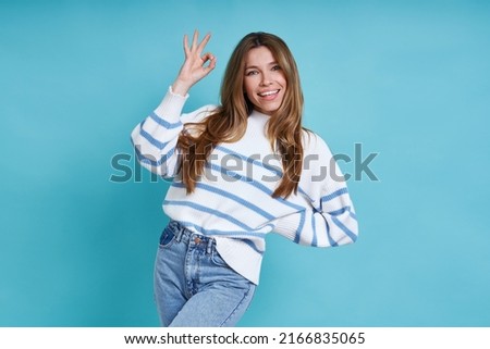 Beautiful young woman looking at camera and gesturing OK sign while standing against blue background