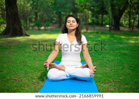 Beautiful young indian woman wearing white cloths doing yoga meditation outdoor in nature Brunette female breathing exercise in park alone, fitness,  Mental health, Peaceful mind,. Copy space.  Royalty-Free Stock Photo #2166859991