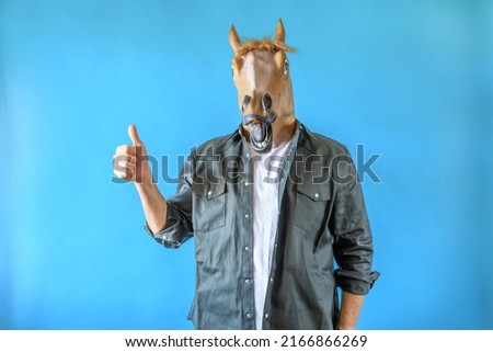 Funny horse head on human body on a green shirt on blue background, thumbs up and looking at camera. Clip art, negative space.
