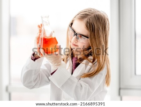 Smart girl during scientific chemistry experiment wearing protection glasses, holding bottle with orange liquid. Schoolgirl with chemical equipment on school lesson portrait