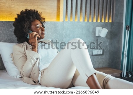 An African business woman on a business trip is lying on a bed and talking on the phone in a hotel room. A woman in formal clothes lies in a hotel room and makes a phone call.