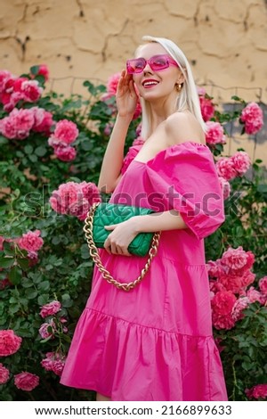 Happy smiling fashionable woman wearing trendy summer fuchsia color dress, pink sunglasses, holding stylish green faux leather bag, posing near blooming roses Royalty-Free Stock Photo #2166899633