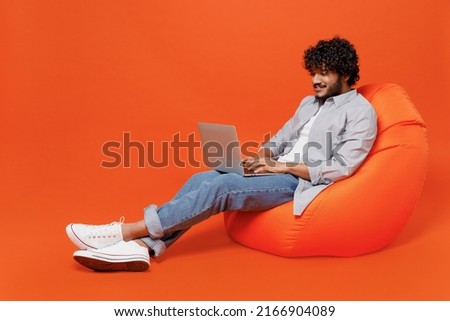 Full size body length young bearded Indian man 20s years old wears blue shirt sit in bag chair hold use work on laptop pc computer typing browsing isolated on plain orange background studio portrait