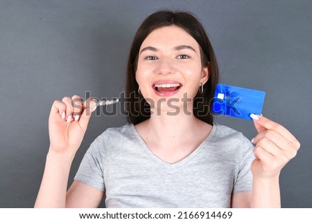Young beautiful caucasian girl standing over gray background holding a credit card and an invisible aligner braces and smiling. Dental healthcare concept. 