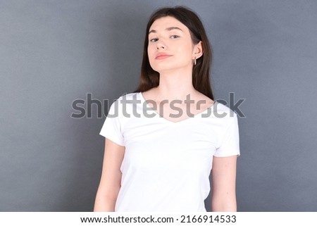 beautiful Caucasian woman wearing white T-shirt over studio grey wall with snobbish expression curving lips and raising eyebrows, looking with doubtful and skeptical expression, suspect and doubt. Royalty-Free Stock Photo #2166914533