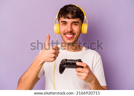 Young hispanic man playing with a video game controller isolated on purple background smiling and raising thumb up