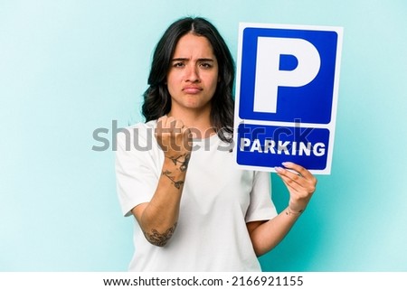Young hispanic woman holding parking placard isolated on blue background showing fist to camera, aggressive facial expression.