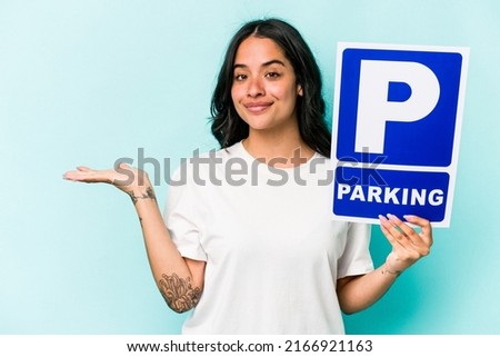 Young hispanic woman holding parking placard isolated on blue background showing a copy space on a palm and holding another hand on waist.